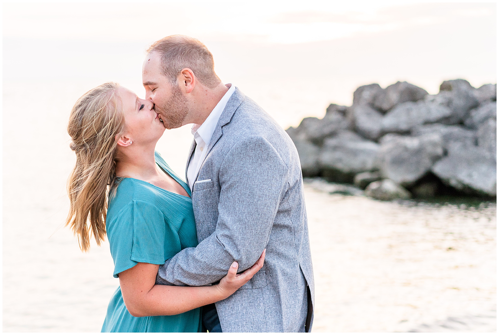 Romantic formal engagement portraits at Sims Park beach in Cleveland Ohio. Woman is wearing a long formal teal gown with flowing sleeves. The man is in a grey suit and white button up shirt. He is holding his fiance by the waist and kissing her, both have their eyes closed. 